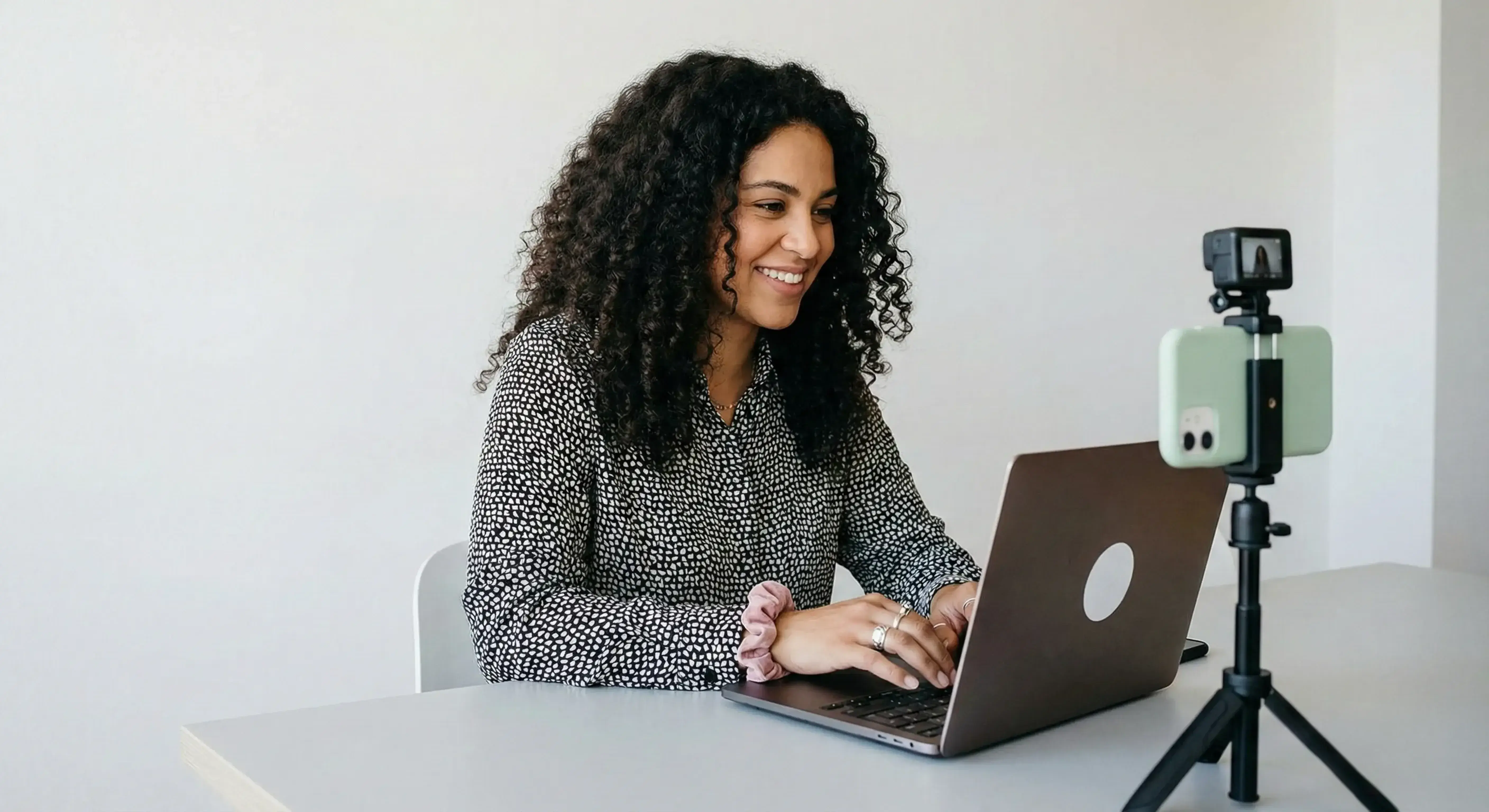 Mulher sentada em uma cadeira de escritório usando um notebook cinza e com um celular em um tripé fazendo uma gravação